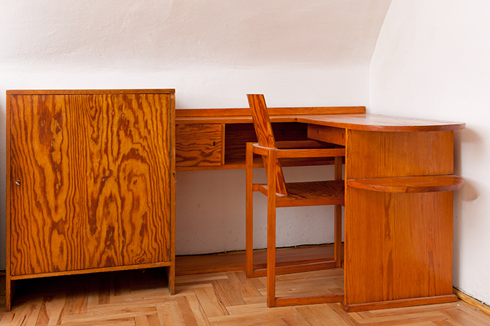 Minimalistyczne meble z jasnego drewna w narożniku: komoda i biurko w kształcie "L" z krzesłem. Wyraźne usłojenie, ciepły odcień. Pomieszczenie z białymi ścianami i parkietem w jodełkę.