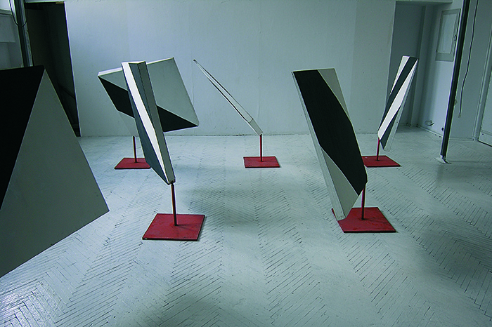 Instalacja artystyczna. Kilka abstrakcyjnych rzeźb z czarno-białych paneli na pomarańczowo-czerwonych stojakach. Rzeźby r&oacute;żnią się kątem nachylenia i ułożeniem kolorowych pas&oacute;w.  Tło jest proste, białe ściany w przestronnej galerii.