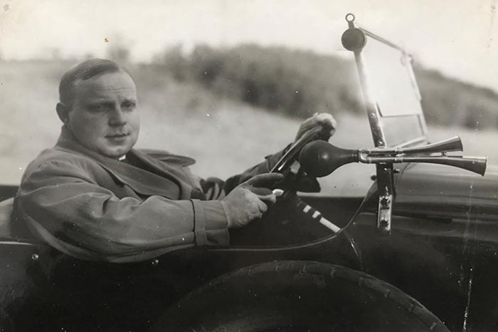 Czarno-białe zdjęcie przedstawia mężczyznę za kierownicą zabytkowego samochodu. Mężczyzna, ubrany w płaszcz, spogląda na wprost, z dłońmi na kierownicy. Samoch&oacute;d ma z boku trąbki klaksonu. 
Tło jest rozmyte, przypomina wiejski krajobraz.