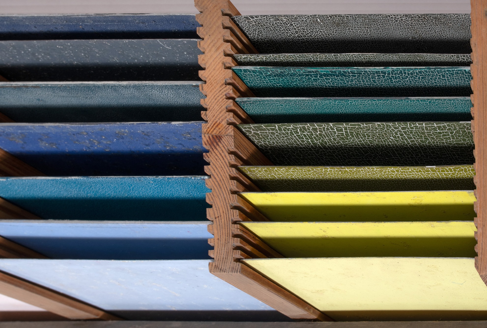 Drewniane p&oacute;łki z pr&oacute;bkami kolor&oacute;w. Po lewej błękity i szarości, po prawej zielenie, szarości (często spękane) i ż&oacute;łcie. Pr&oacute;bki ułożone poziomo, ukazując barwy i teksturę.